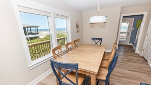 Comedor con mesa de madera y sillas azules, ventanas amplias con vista a la playa y un pasillo que conduce a otra habitación.