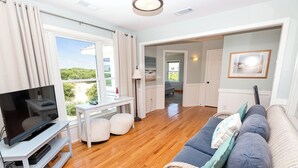 A living room with a television, white furniture, a wicker sofa, and a view through a large window. - Oceanside, Private Pool, Hot Tub, ELEV, Close to Beach Access, OS1C, Here At Last (Corolla)