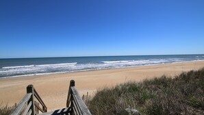 Ein hölzerner Steg führt zu einem Sandstrand, im Hintergrund ist das Meer zu sehen.