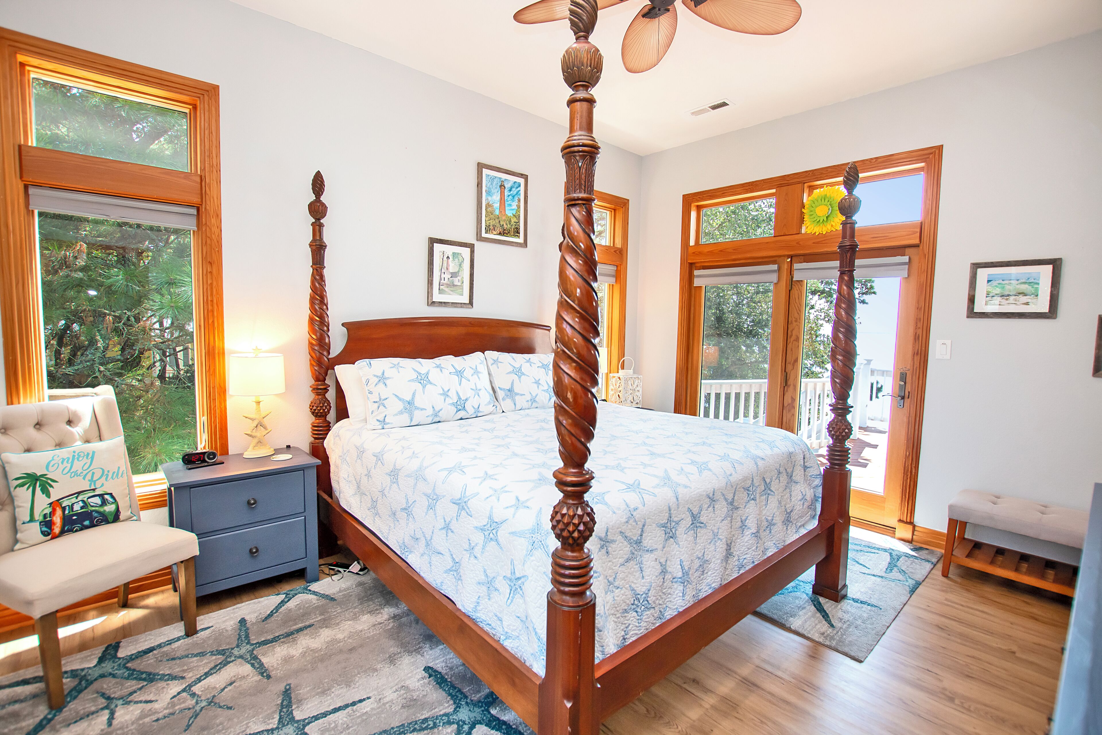 A four-poster bed with a star-patterned bedspread, a wooden nightstand with a lamp, a bench, and a view of the outdoors through large windows.
