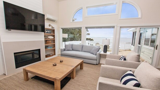 Un salón amplio con una televisión grande, una chimenea, un sofá y una mesa de centro. Varias ventanas ofrecen vistas al exterior.