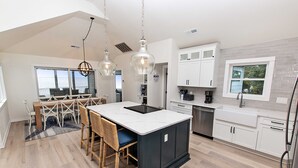 Una cocina moderna con una isla central, armarios blancos y un comedor con sillas de mimbre.