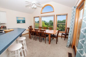 A dining area with a wooden table and chairs, a kitchen counter with stools, and large windows offering a view of the outdoors.