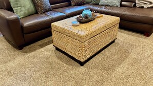 Un sofá seccional de cuero marrón con varios cojines, una mesa de centro tejida con objetos decorativos y una alfombra color beige.