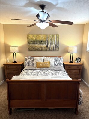 Une chambre à coucher avec un lit en bois, deux tables de chevet, un ventilateur de plafond, un tableau représentant une forêt et un coussin orné du mot « ensemble ».