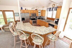 A kitchen with wooden cabinets, a granite countertop, stainless steel appliances, and a bar area with chairs. - Oceanside, Rec Room, Priv Pool, Hot Tub, Close to Beach Access, PI161, St Nicks (Corolla)