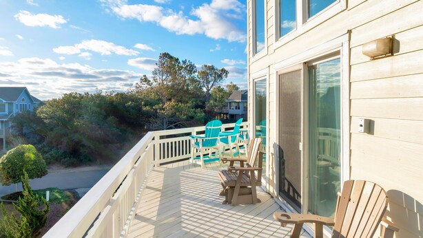 A wooden deck with outdoor furniture, including chairs and a bench, overlooking a coastal area with trees and houses.