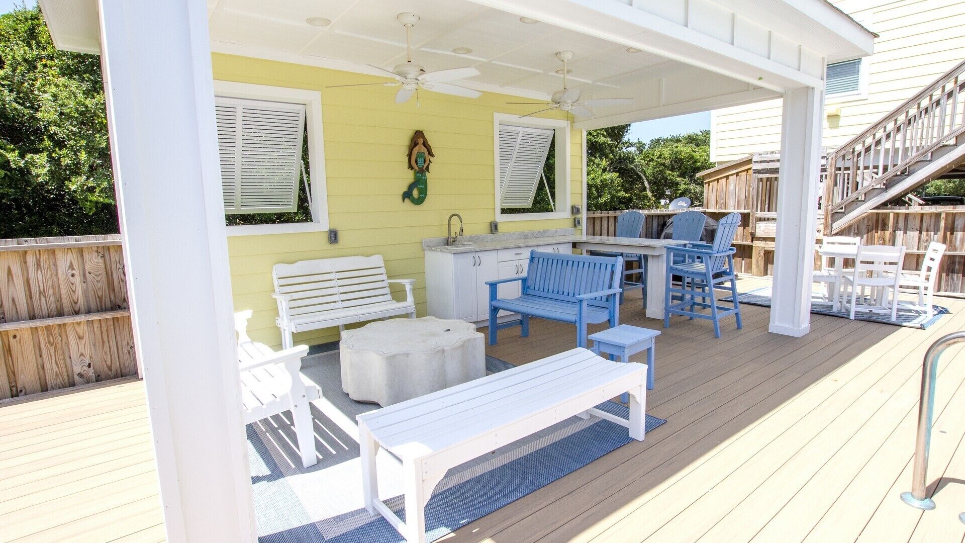 Un patio techado al aire libre con zona de comedor, mobiliario blanco y un ventilador de techo.