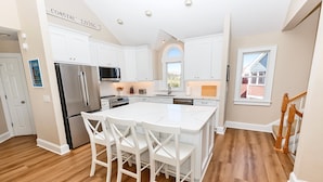 A spacious kitchen with white cabinetry, a large island with bar stools, stainless steel appliances, and a view of the outdoors through a window. - Oceanside, Private Pool, Hot Tub, Buck Island Amenities, BU21, Island Girl (Corolla)