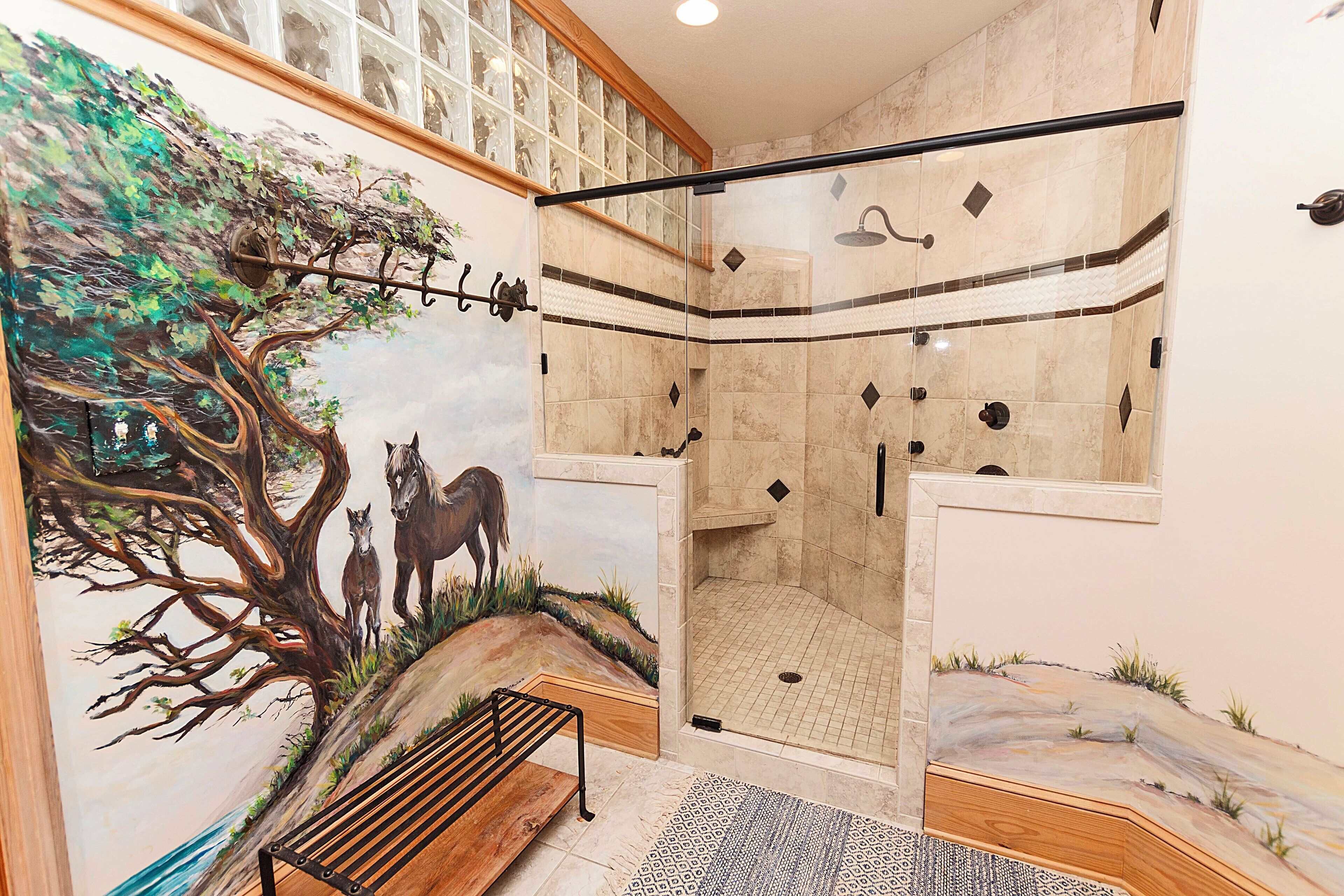 Un espace douche doté d’une porte en verre, d’un banc et d’une fresque représentant un arbre et des chevaux.