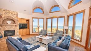 A spacious living room with a stone fireplace, comfortable seating, large windows with ocean views, and a television. - OCEANFRONT, 9 BRs, Private Pool, Theater Rm, Rec Rm, Ocean Views, PI134, Heavenly Dayz (Corolla)