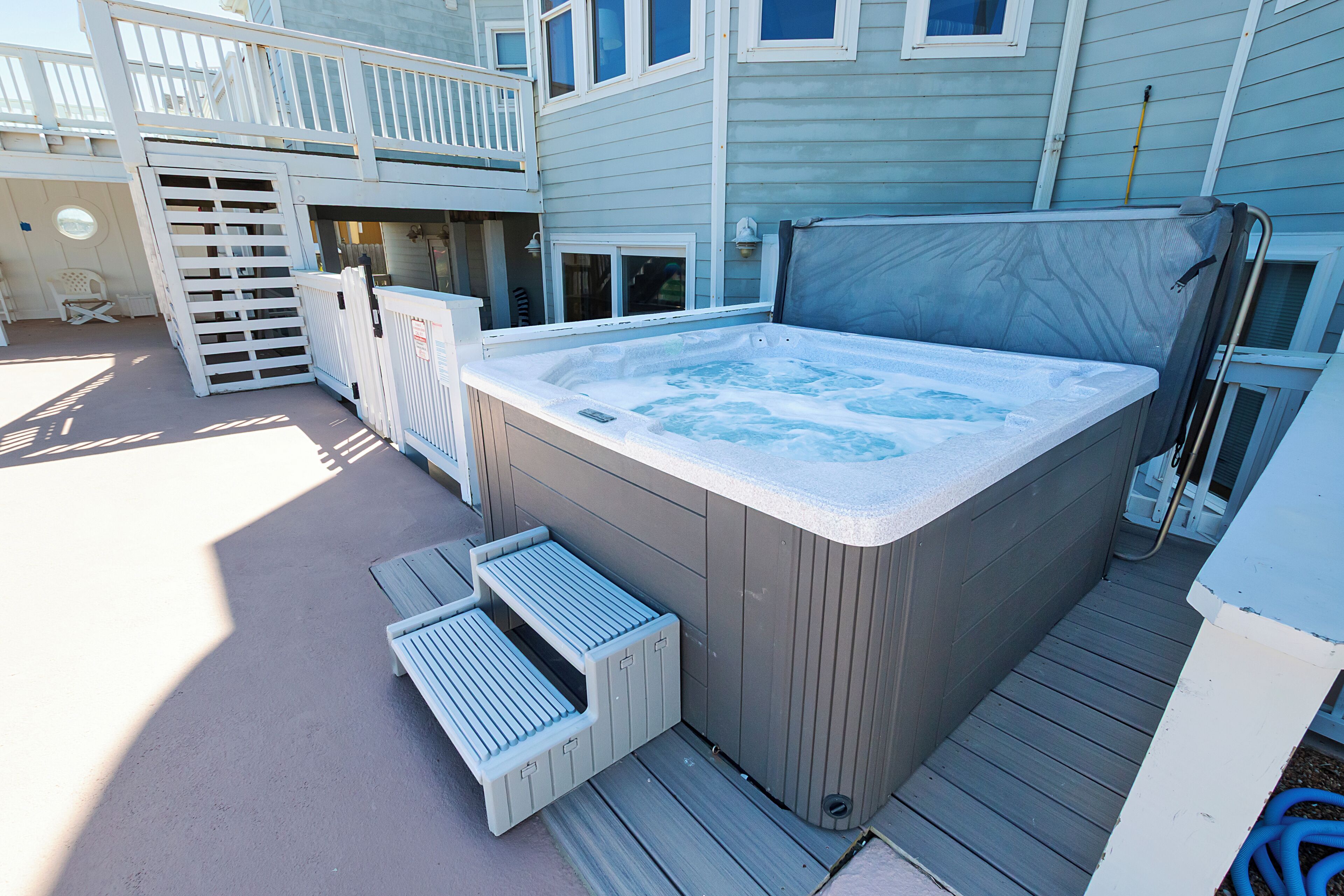 Un bain à remous installé sur une terrasse, accessible par des marches, entouré d’une terrasse en bois et d’une maison dotée d’un balcon.
