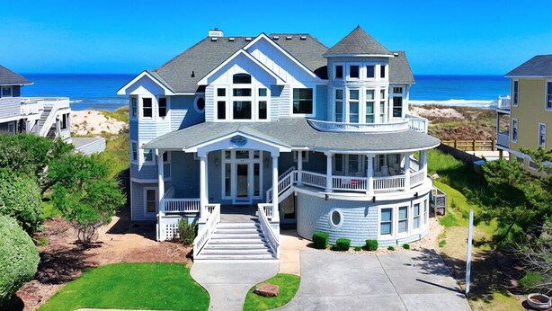A large, two-story house with a wrap-around porch and a balcony, situated near a beach. - OCEANFRONT, 9 BRs, Private Pool, Theater Rm, Rec Rm, Ocean Views, PI134, Heavenly Dayz (Corolla)