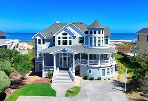 A large, two-story house with a wrap-around porch and a balcony, situated near a beach.