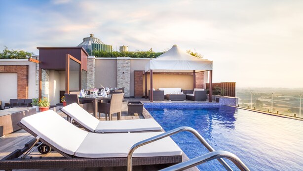 An infinity pool with lounge chairs and a dining area, surrounded by modern architecture and a view of the city. - Hotel Almeida (Dera Bassi)