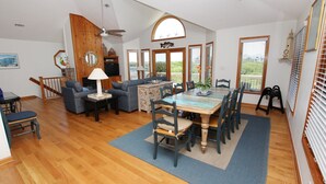 A spacious living room with a wooden floor, a dining area with a large table and chairs, a sofa set, and a ceiling fan. - Oceanside, Hot Tub, Screened porch, Close to Beach, OS9H, Starfisher (Corolla)