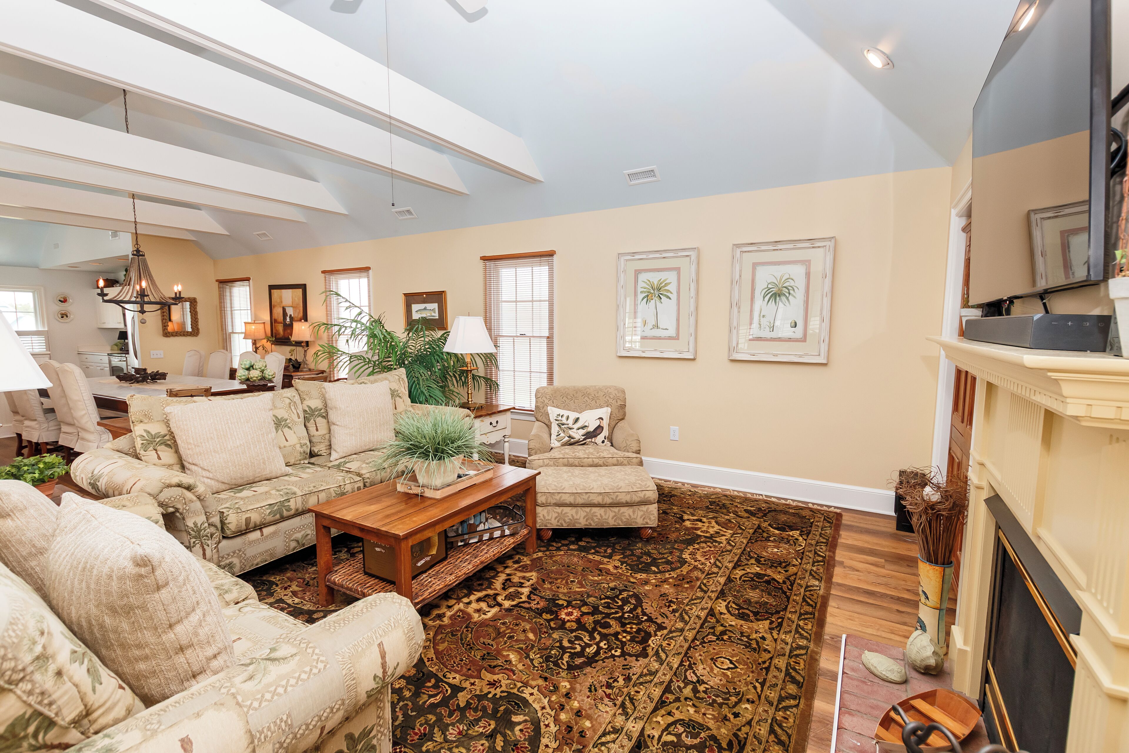A spacious living room with a large sofa, a coffee table, and a fireplace. There are framed pictures on the wall and a television mounted above the fireplace.
