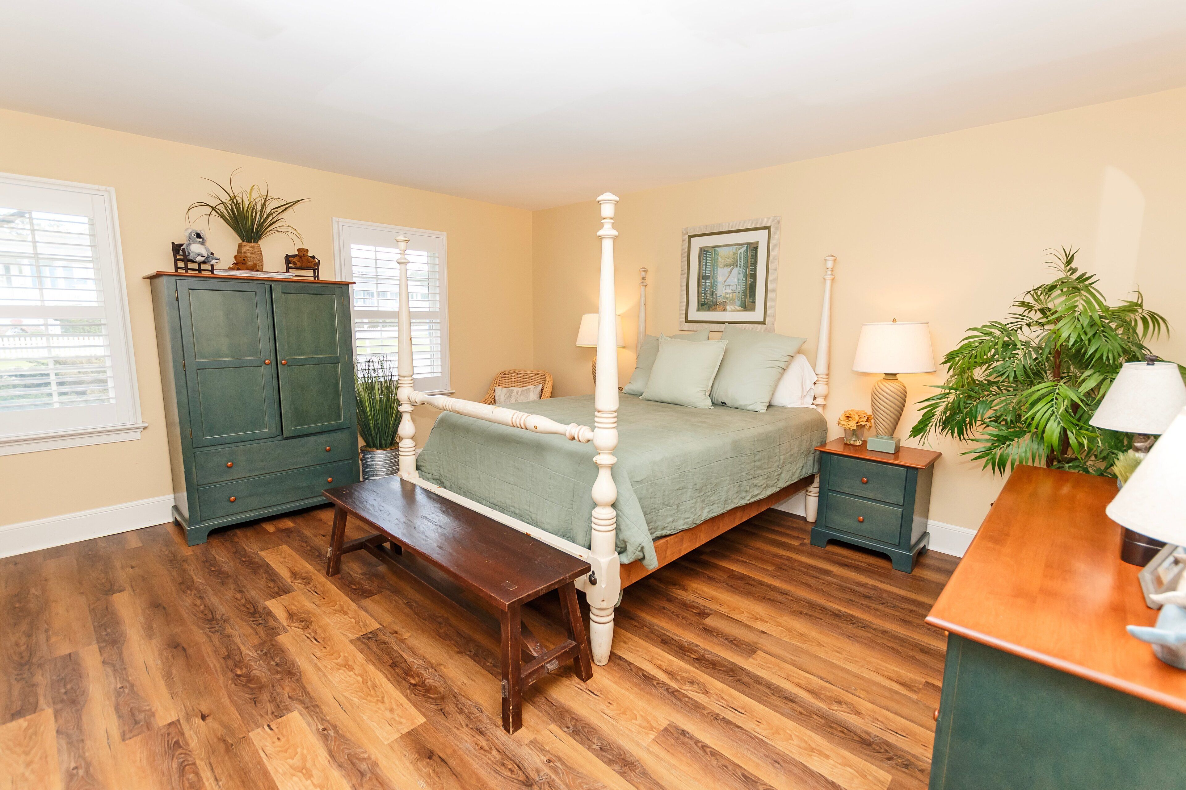 Quarto com cama de dossel, piso de madeira, penteadeira verde, mesa de cabeceira e janela com persianas.
