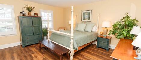 Quarto com cama de dossel, piso de madeira, penteadeira verde, mesa de cabeceira e janela com persianas.