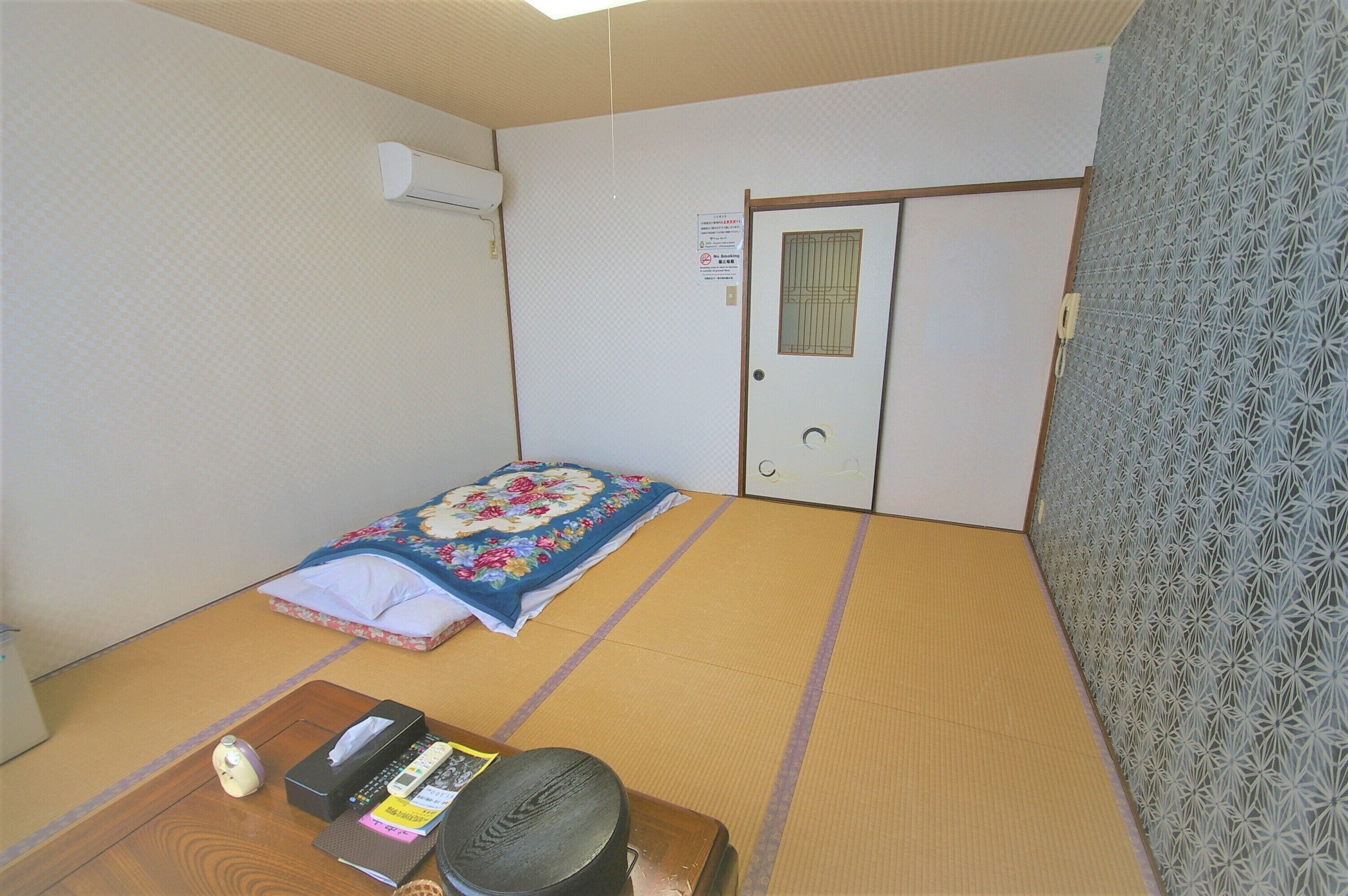 A traditional Japanese room with tatami flooring, a futon bed, a wooden desk with a telephone, and a patterned wall.