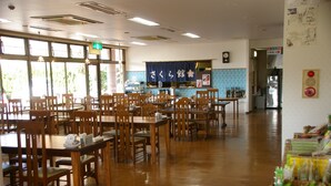 A spacious dining area with wooden tables and chairs, a kitchen in the background, and a display of food items on the right.