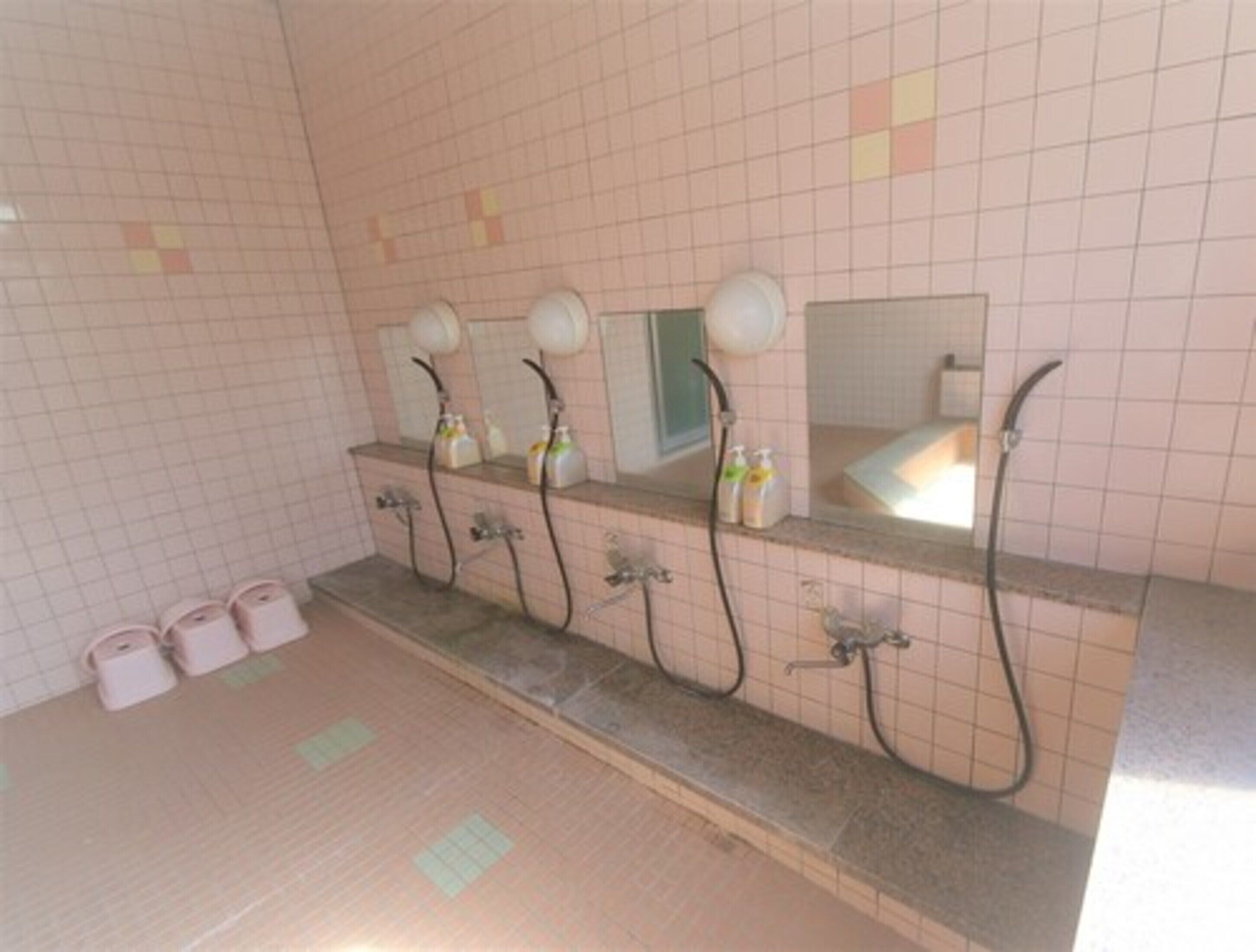 Baño con cuatro lavamanos, cada uno con espejo y cabezal de ducha manual. Hay tres rollos de papel higiénico en el suelo.