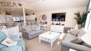 A spacious living room with a grey sofa, teal armchairs, and a white coffee table. A kitchen area with white cabinets and a dining table is visible in the background. - Oceanside, Dogs Welcome, Hot Tub, Close to Shopping, BU13A, Tropical Dreams A (Corolla)