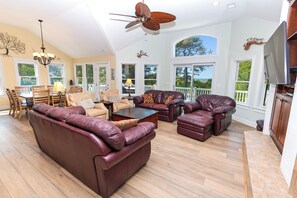 A spacious living area with leather sofas, a dining table, and chairs. There is a ceiling fan and large windows offering views of the outdoors.