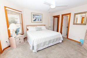 A bedroom with a bed, a ceiling fan, a window with blinds, a bedside table with a lamp, and a framed picture on the wall.