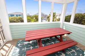 Un patio couvert avec une table de pique-nique rouge et des bancs, une vue sur les maisons à travers de grandes fenêtres et un tapis à motifs sur le sol.