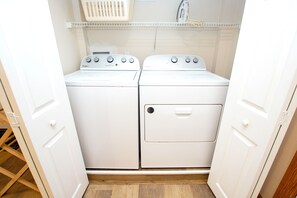 A laundry room with two white Whirlpool washers and dryers, a white shelf above, and a wooden floor. - Oceanside, Hot Tub, Pool Table, Community Pool, CP2, The Green Wave (Corolla)