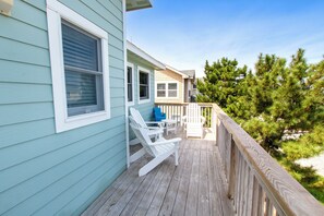 A wooden deck with white chairs and a table, a light blue building with a window, and a pine tree in the background. - Oceanside, Hot Tub, Pool Table, Community Pool, CP2, The Green Wave (Corolla)