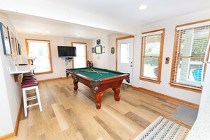 Une table de billard dans une pièce dont le sol est recouvert de parquet, avec un téléviseur fixé au mur et une porte dotée d’une fenêtre.