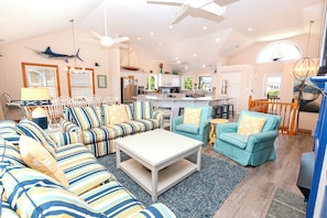 A spacious living room with a large sofa set, a coffee table, and a kitchen area with white cabinets and a central island. - Oceanside, Hot Tub, Pool Table, Community Pool, CP2, The Green Wave (Corolla)