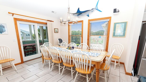 Un coin repas avec une table en bois et des chaises blanches, un lustre et une vue par les fenêtres.