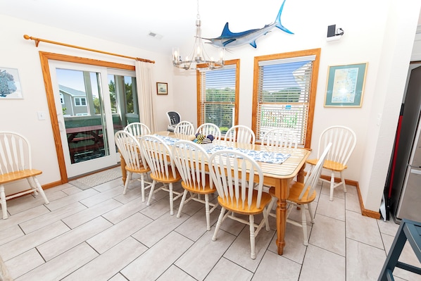 A dining area with a wooden table and white chairs, a chandelier, and a view through the windows. - Oceanside, Hot Tub, Pool Table, Community Pool, CP2, The Green Wave (Corolla)