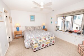 A bedroom with a bed, two bedside tables, a window with a view, a ceiling fan, and a small window sill with decorative items.
