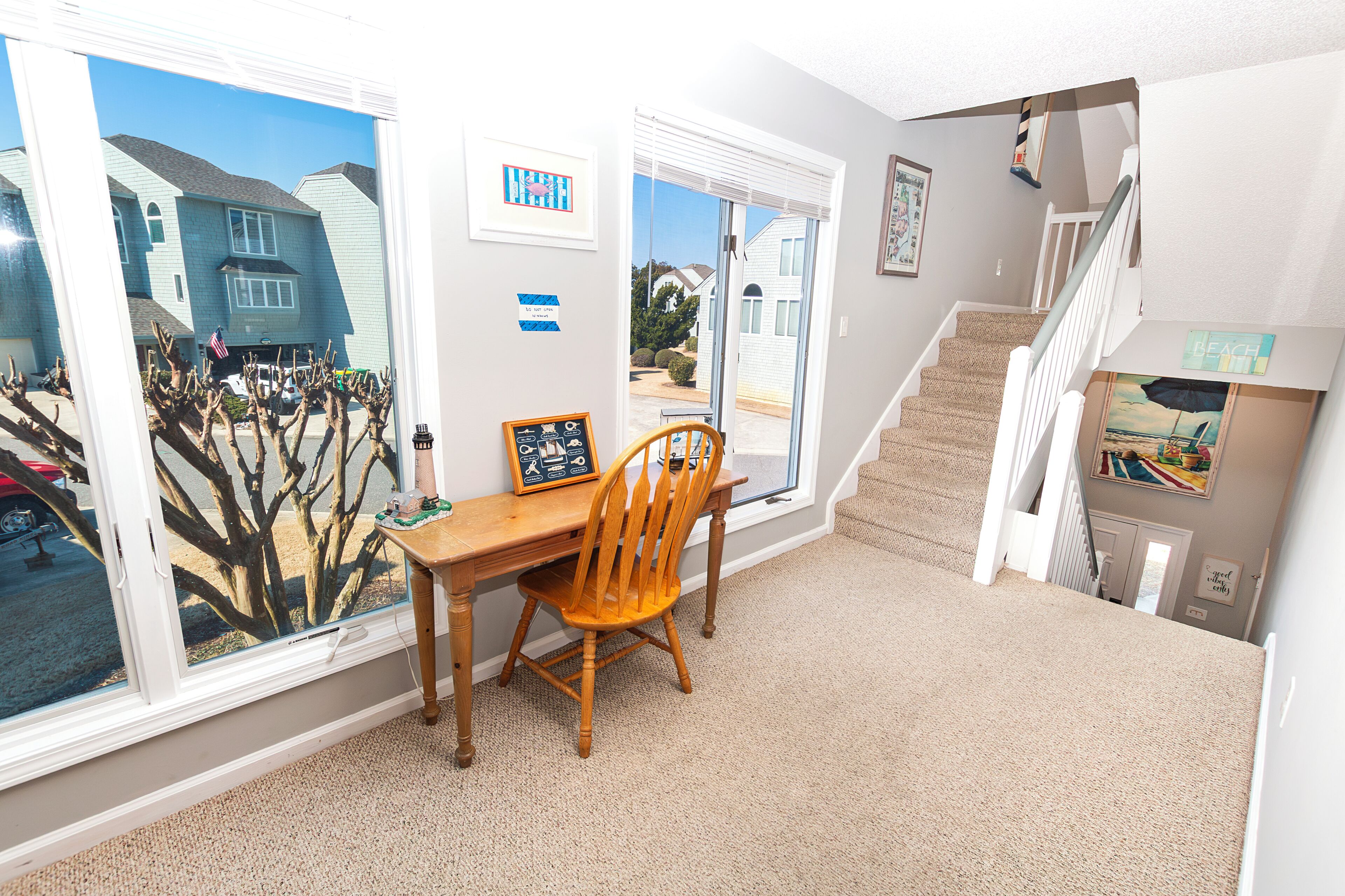 A room with a wooden desk, a chair, a staircase, and a view of a house through a window.