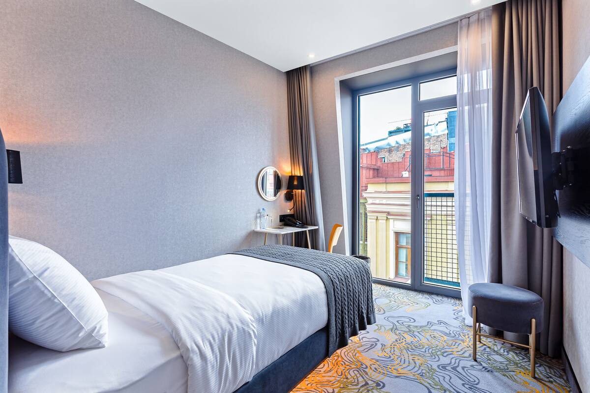A hotel room with a bed, a small table with a lamp, a TV, and a view of the outside through a large window.