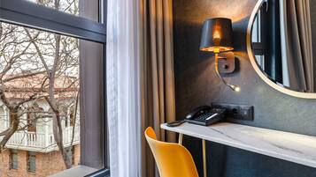 A hotel room with a large window, a desk with a telephone, a chair, and a wall-mounted lamp.