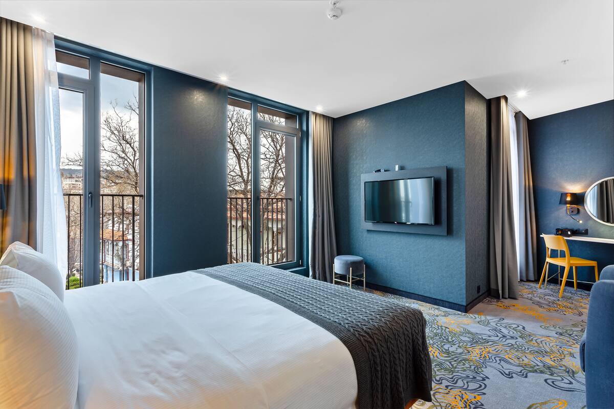 A hotel room with a large bed, a desk with a chair, a TV mounted on the wall, and a balcony with a view of trees and buildings.