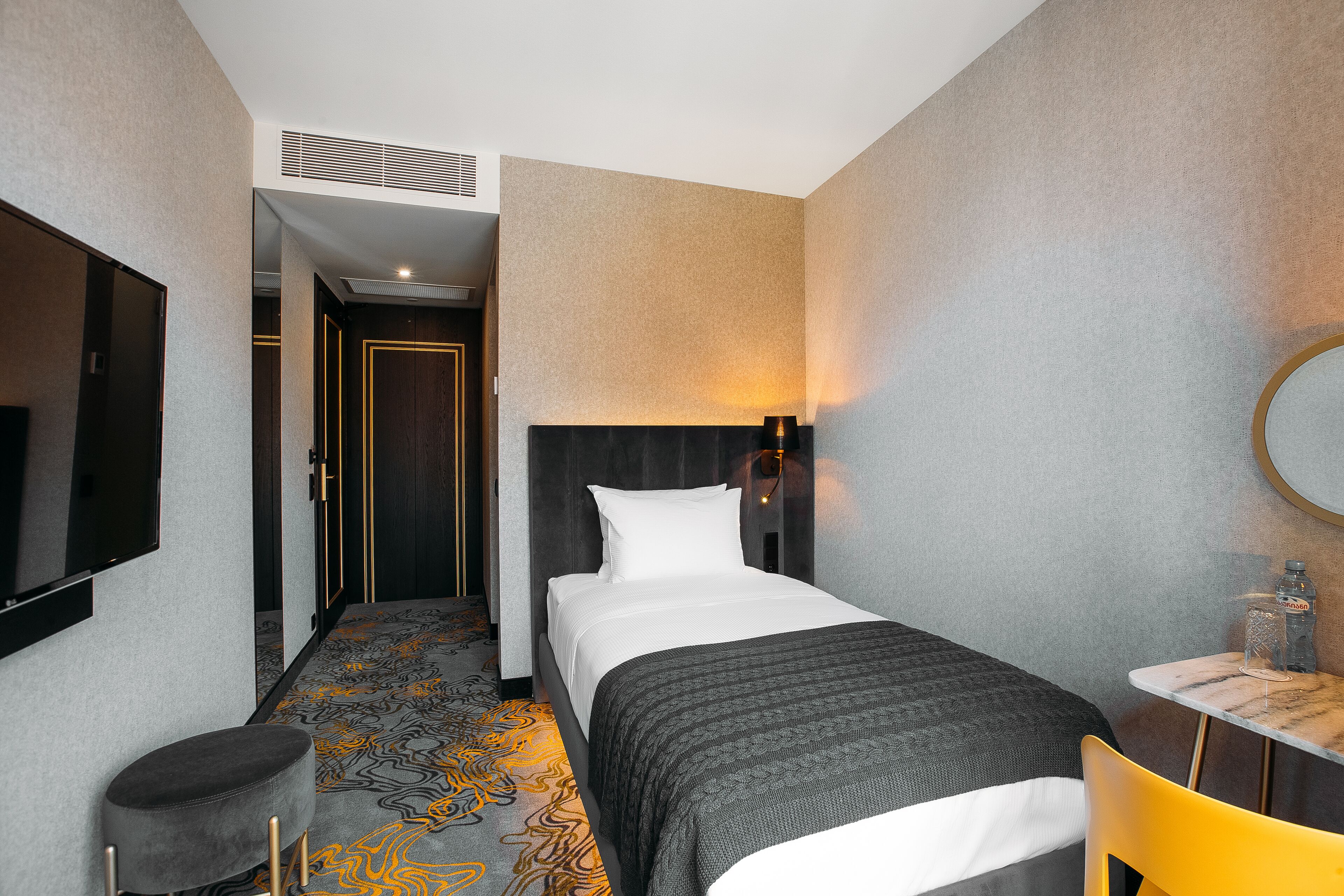 A hotel room with a bed, a TV, a small table with a bottle, a chair, and a patterned carpet.