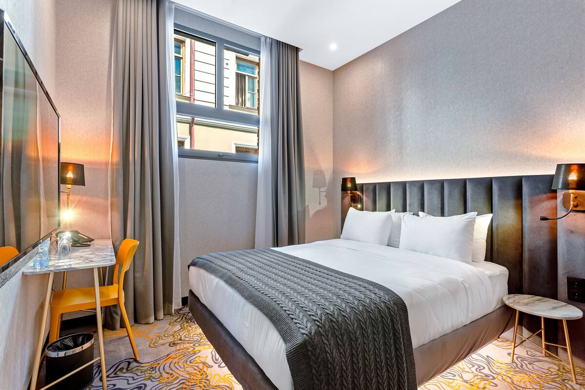 A hotel room with a large bed, a desk with a chair, a small round table, and a trash bin.