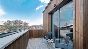 Een balkon met een houten vloer, twee stoelen, een klein tafeltje en een bed met uitzicht op de stad.