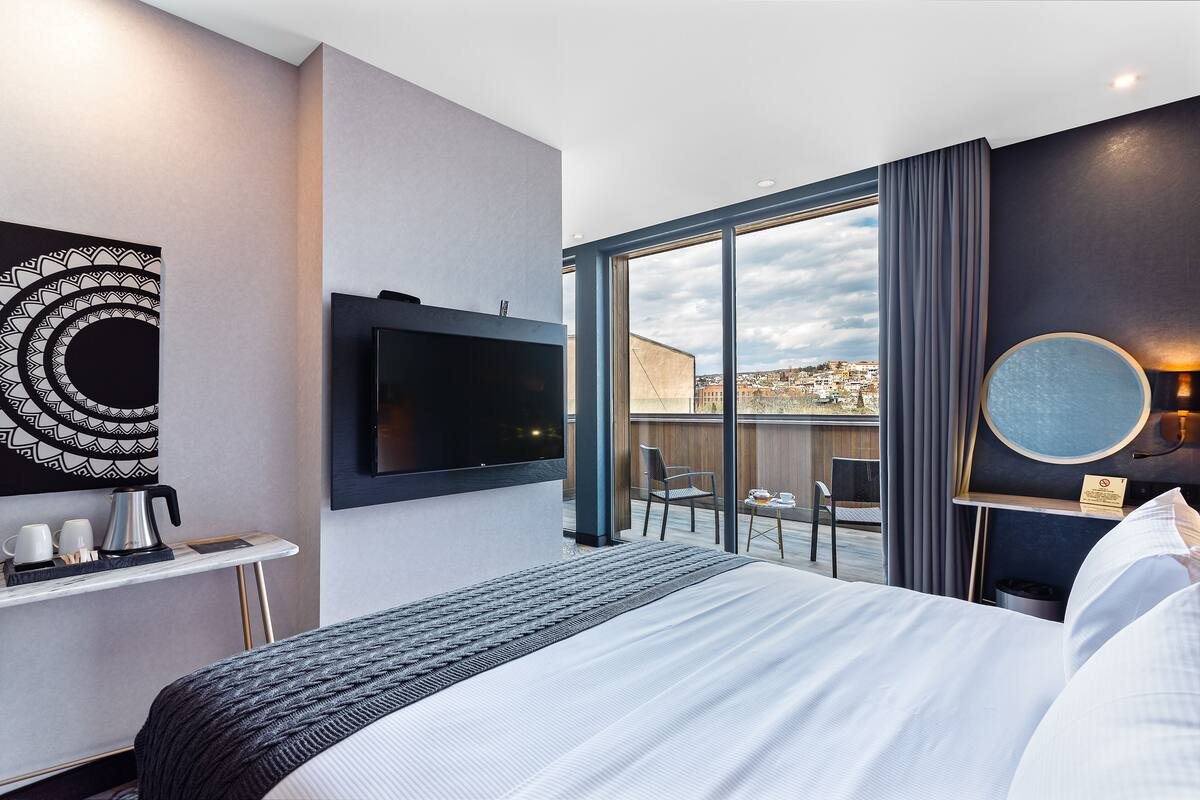 A modern hotel room with a large bed, a flat-screen TV, a coffee station, and a balcony with a city view.