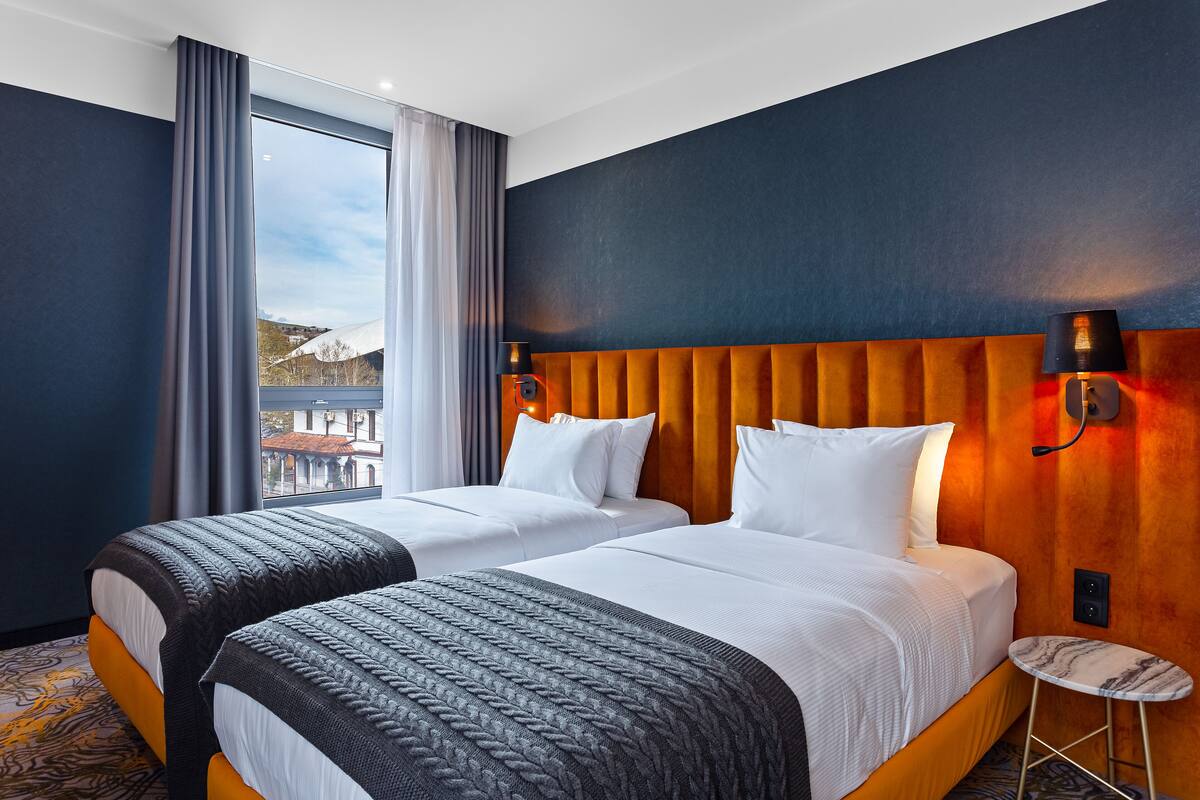 A hotel room with two beds, a large window with curtains, a wooden headboard, and a small round table.