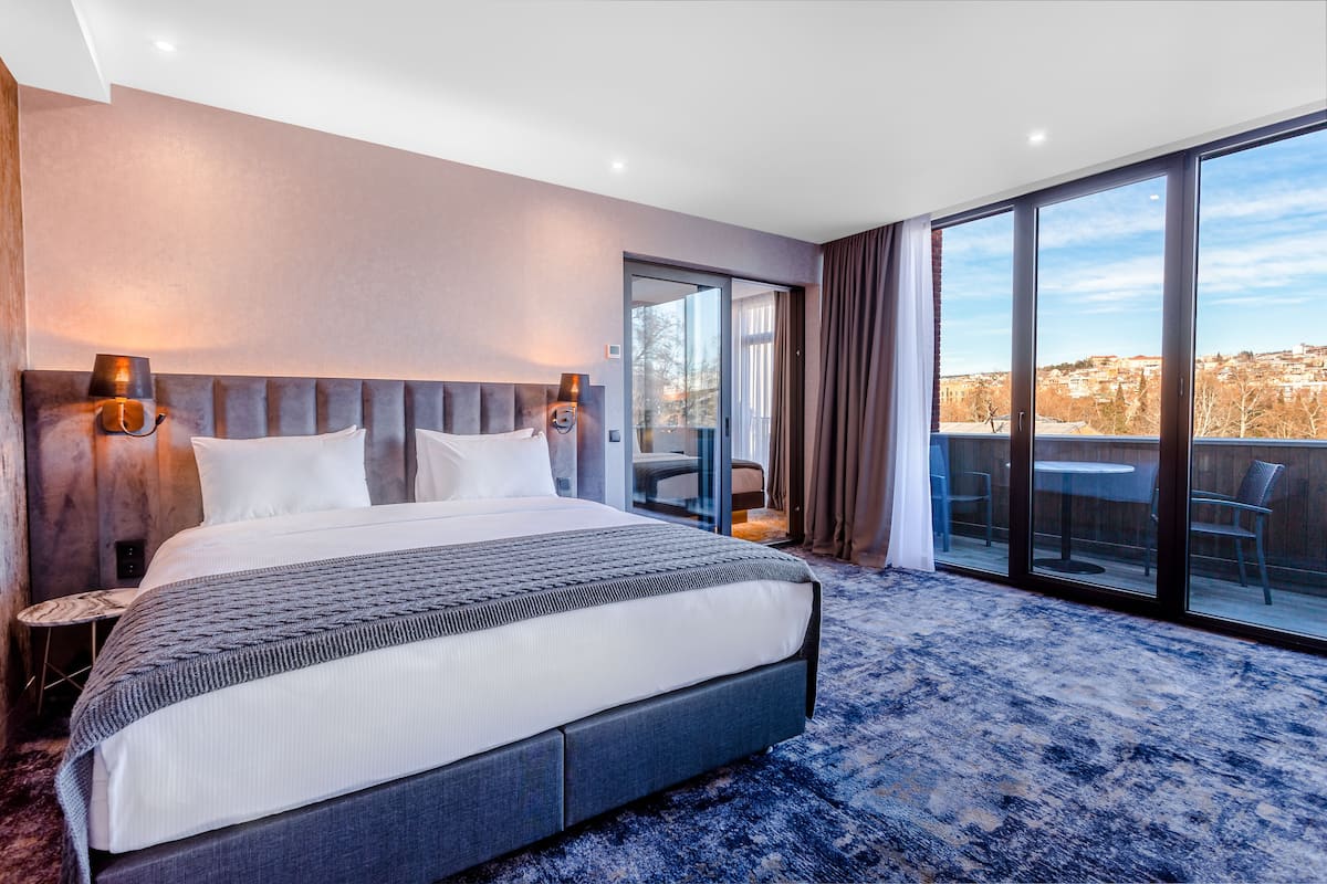 A modern hotel room with a large bed, a glass door leading to a balcony, and a city view.