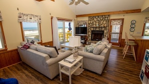 A cozy living room with a stone fireplace, wooden flooring, and a view of the beach through large windows. - OCEANFRONT, 4-wheel drive area, Wild Horses, Ocean Views! 4x1662, Dolphin Watch (Corolla)