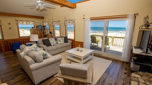 A cozy living room with a wooden floor, a beige sofa, a white coffee table, and a view of the beach through large sliding glass doors. - OCEANFRONT, 4-wheel drive area, Wild Horses, Ocean Views! 4x1662, Dolphin Watch (Corolla)