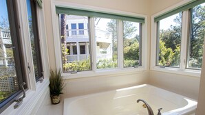 A bathroom with a large bathtub, a window with a view of a house and trees, and a potted plant on the windowsill. - Soundside, Community Amenities, Sun Decks, 327, Footprints by the Sound (Corolla)